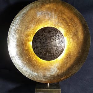 May include: A large, circular, gold-coloured metal sculpture with a textured surface. The sculpture has a dark, circular centre and is mounted on a square base. The interior glows with a warm light.