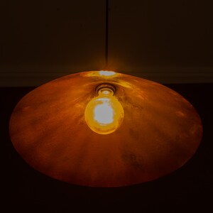 Op de afbeelding: Een koperkleurige hanglamp met een enkele zichtbare gloeilamp. De lamp heeft een grote, afgeronde kap en hangt aan een dunne draad. De lamp brandt en geeft een warme gloed.