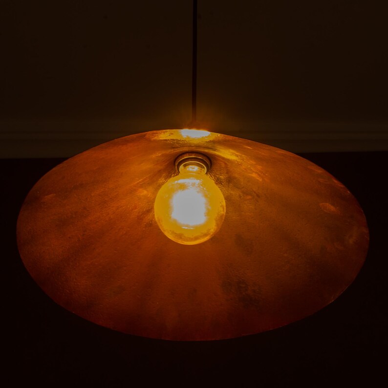 Op de afbeelding: Een koperkleurige hanglamp met een enkele zichtbare gloeilamp. De lamp heeft een grote, afgeronde kap en hangt aan een dunne draad. De lamp brandt en geeft een warme gloed.