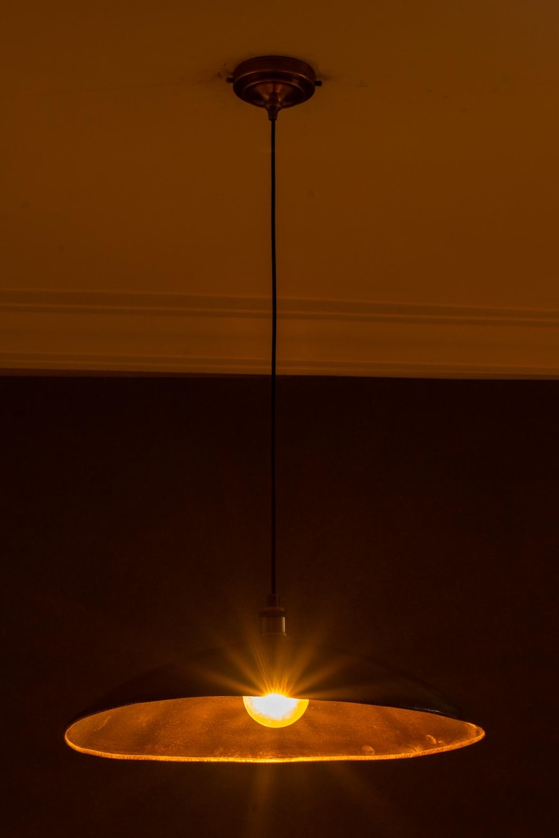 May include: A pendant light fixture with a dark, metallic shade and a bright, exposed bulb. The light is suspended from a dark cord attached to a circular ceiling mount. The overall color scheme is warm, with shades of brown and gold.
