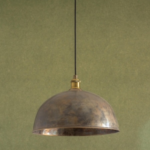Op de afbeelding: Een hanglamp met een koepelvormige kap met een bronzen afwerking en messing fitting. Het licht hangt aan een zwart snoer. De achtergrond is groen. De lamp heeft een vintage of industriële stijl.