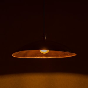 May include: A hanging, dark-colored pendant light with a wide, shallow shade. The interior of the shade has a warm, golden hue, and a single light bulb is visible, emitting a soft, yellow glow. The background is a deep, dark brown.