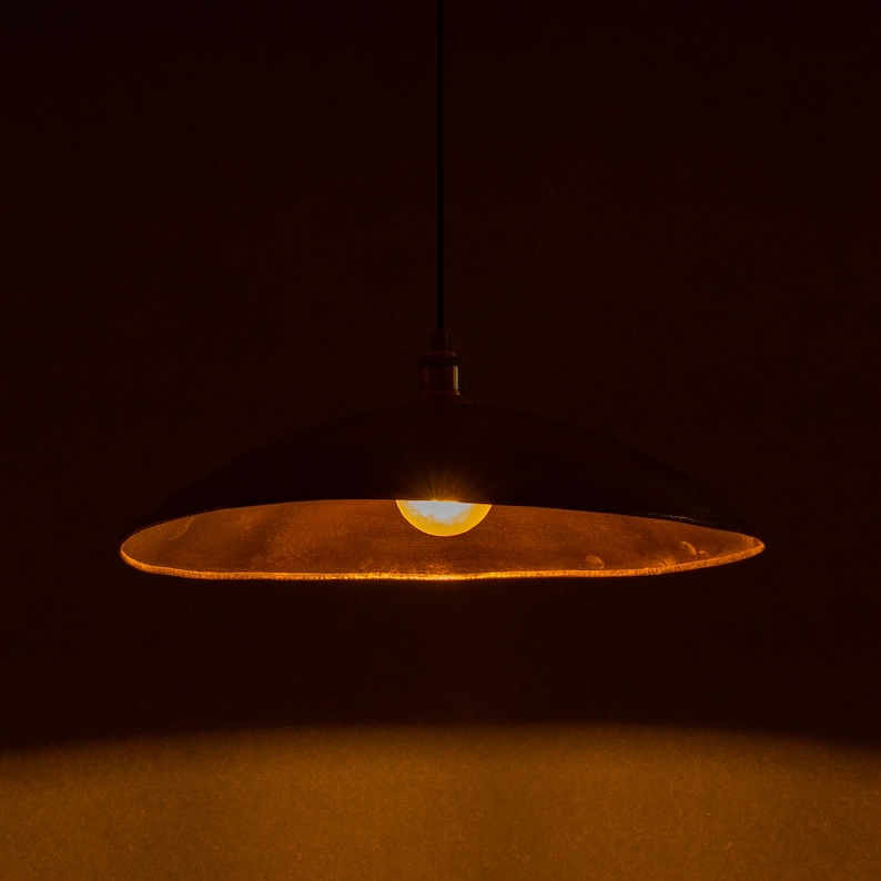 May include: A hanging, dark-colored pendant light with a wide, shallow shade. The interior of the shade has a warm, golden hue, and a single light bulb is visible, emitting a soft, yellow glow. The background is a deep, dark brown.