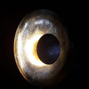 May include: A large, circular, brass-coloured gong with a dark, circular cutout in the centre. The gong has a textured surface and is illuminated from behind, creating a bright, glowing effect against a black background.