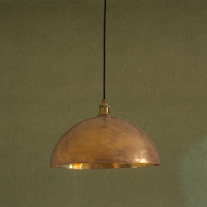 Op de afbeelding: Een messing hanglamp met een koepelvormige kap. De kap heeft een warme, gouden tint en een licht getextureerd oppervlak. De lamp hangt aan een zwart snoer tegen een gedempte groene achtergrond.