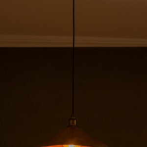Op de afbeelding: Een hanglamp met een koperkleurige, conische kap. De gloeilamp is zichtbaar en geeft een warme gloed. De lamp hangt aan het plafond met een zwart snoer en een messingkleurige kap.