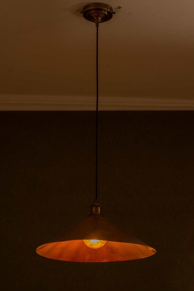 Op de afbeelding: Een hanglamp met een koperkleurige, conische kap. De gloeilamp is zichtbaar en geeft een warme gloed. De lamp hangt aan het plafond met een zwart snoer en een messingkleurige kap.