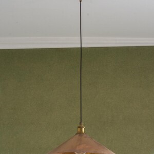 Op de afbeelding: Een koperkleurige hanglamp met een conische kap en een messingkleurige plafondbevestiging. Het licht hangt aan een zwart snoer tegen een groene muur. De armatuur is in vintage- of industri&euml;le stijl.