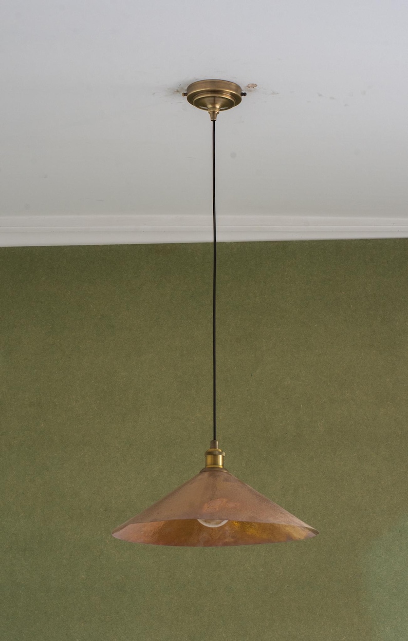 Op de afbeelding: Een koperkleurige hanglamp met een conische kap en een messingkleurige plafondbevestiging. Het licht hangt aan een zwart snoer tegen een groene muur. De armatuur is in vintage- of industri&euml;le stijl.