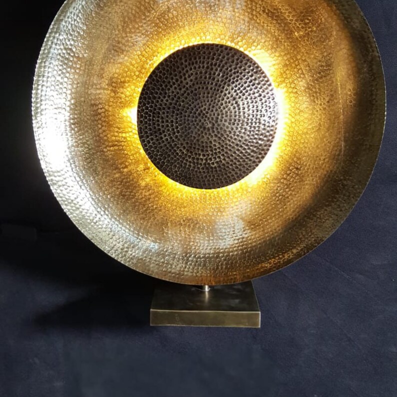 May include: A gold-coloured metal sculpture with a circular design. The sculpture has a textured surface and a dark, circular centre. It is mounted on a square base. The interior is illuminated with a warm, golden light.