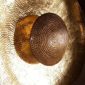 May include: A close-up shot of a golden gong with a textured surface and a smaller, darker brown disc in the centre. The gong's hammered texture creates a reflective surface. The lighting accentuates the metallic sheen and intricate details.