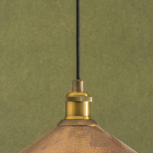 Op de afbeelding: Close-up van een messing hanglamp met een getextureerde, koperkleurige kap. De lamp heeft een zwart snoer en een messing fitting. De achtergrond is een gedempt groen.