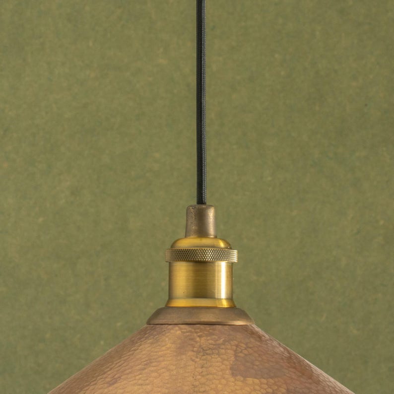Op de afbeelding: Close-up van een messing hanglamp met een getextureerde, koperkleurige kap. De lamp heeft een zwart snoer en een messing fitting. De achtergrond is een gedempt groen.