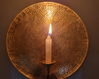 May include: A lit white candle in a brass candle holder, set against a textured, hammered brass plate. The plate is round and mounted on a wall. The candle flame glows brightly in a dimly lit room.