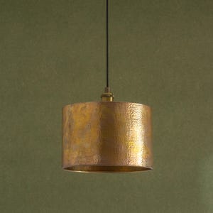 May include: A cylindrical pendant light with a hammered gold finish. It hangs from a black cord and has a brass-coloured fixture at the top. The background is a muted green.