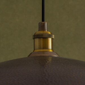 May include: Close-up of a bronze-colored pendant light fixture. The fixture has a textured, hammered metal shade and a brass-colored socket. A black cord suspends the light from above, creating a vintage aesthetic.