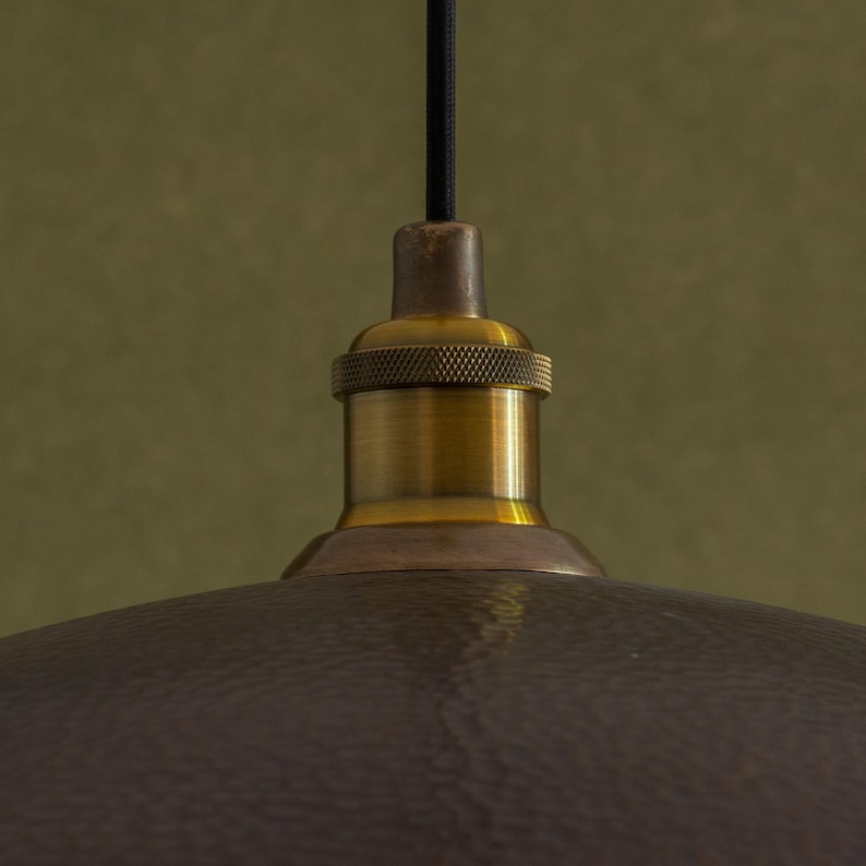 May include: Close-up of a bronze-colored pendant light fixture. The fixture has a textured, hammered metal shade and a brass-colored socket. A black cord suspends the light from above, creating a vintage aesthetic.