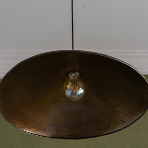 May include: A large, antique brass-colored pendant light fixture. The light has a wide, shallow bowl shape with a textured surface. A single Edison-style bulb hangs from the center, suspended by a black cord. The fixture is designed to be mounted on a ceiling.