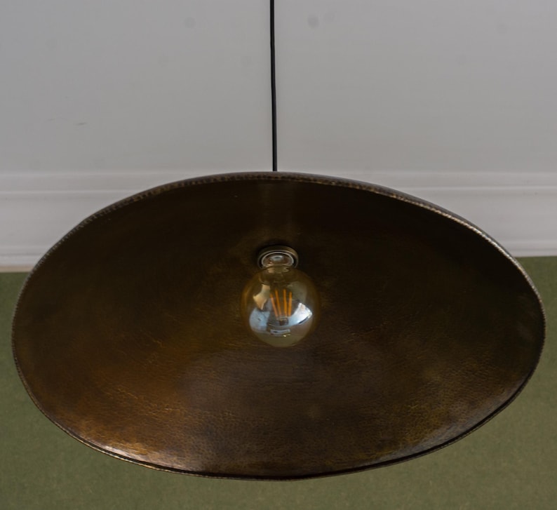 May include: A large, antique brass-colored pendant light fixture. The light has a wide, shallow bowl shape with a textured surface. A single Edison-style bulb hangs from the center, suspended by a black cord. The fixture is designed to be mounted on a ceiling.