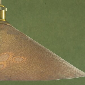Op de afbeelding: Close-up van een hanglamp in vintage stijl. De lampenkap heeft een gehamerde koperkleur met een textuur. De armatuur heeft een messingkleurige bovenkant en een groene achtergrond.