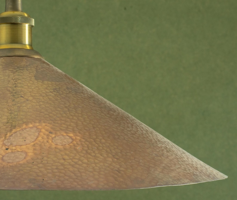 Op de afbeelding: Close-up van een hanglamp in vintage stijl. De lampenkap heeft een gehamerde koperkleur met een textuur. De armatuur heeft een messingkleurige bovenkant en een groene achtergrond.