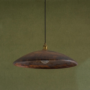 May include: A brown, rustic pendant light with a wide, shallow shade. The shade has a textured, aged appearance. The light hangs from a black cord with a brass-coloured fixture. The bulb is visible.