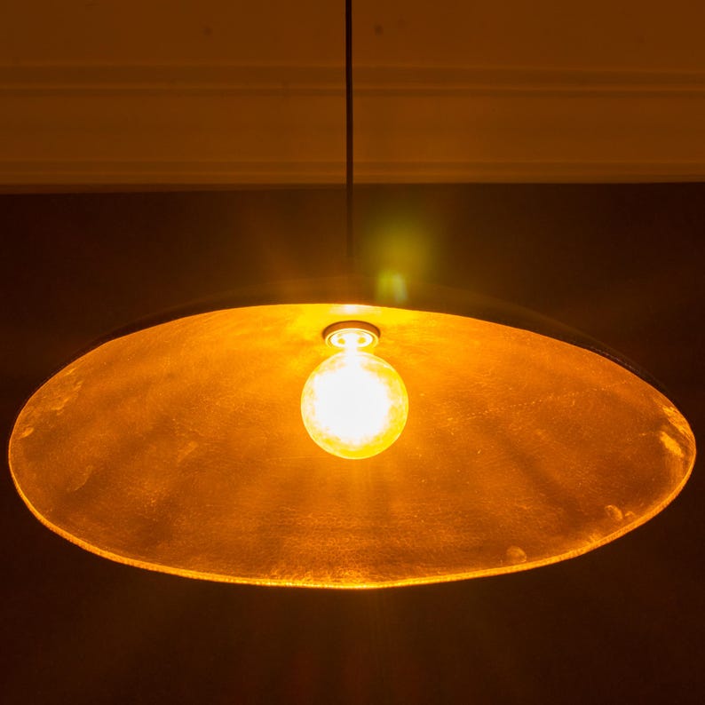 May include: A hanging pendant light with a large, shallow, gold-colored shade. A single, round, illuminated light bulb hangs in the center. The light fixture is suspended from a thin black cord against a dark background.