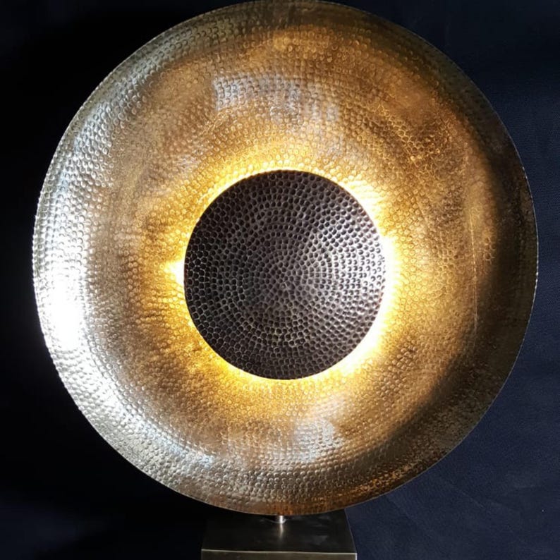 May include: A large, hammered metal sculpture with a circular design. The outer ring is gold, and the centre is dark grey. The sculpture is mounted on a square base. The texture of the metal is visible throughout the piece.