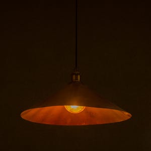 Op de afbeelding: Een hanglamp in vintage-stijl met een conische, bronskleurige kap. De binnenkant van de kap gloeit met warm licht van een centrale lamp. De armatuur hangt aan een zwart snoer tegen een donkere achtergrond.