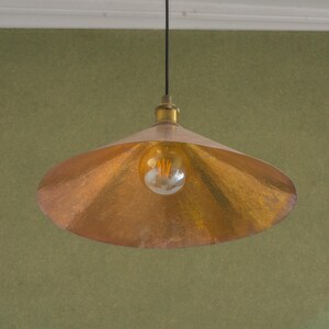Op de afbeelding: Een koperkleurige hanglamp met een getextureerde, komvormige kap. De lamp heeft een messingkleurige fitting en een zichtbare Edison-stijl lamp. De armatuur hangt aan een zwart snoer tegen een groene muur.