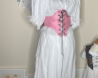 May include: A white peasant top with a pink corset belt. The top has a gathered neckline and lace trim on the sleeves. The corset belt has a lace-up front and is tied at the back.
