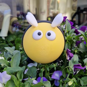 Könnte beinhalten: Gelbe, bienenartige Gartenfigur mit großen Kulleraugen und weißen Antennen. Die Figur ist umgeben von üppigem, grünem Laub und violetten und weißen Stiefmütterchen, was eine fröhliche, dekorative Szene ergibt.