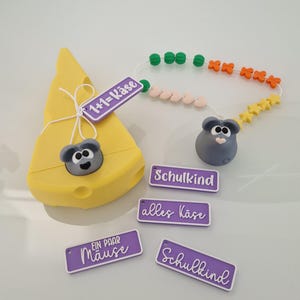 Könnte beinhalten: Ein gelbes Käse-Spielzeug mit einer grauen Maus, begleitet von einer Perlenkette und mehreren lila Etiketten mit deutschem Text. Die Etiketten lauten "1+1=Käse", "Schulkind", "alles Käse" und "Ein Paar Mäuse".