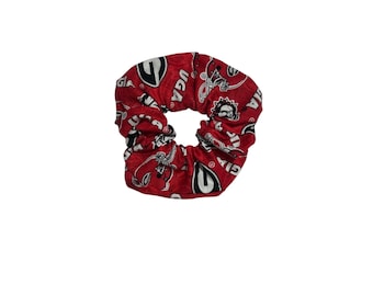 May include: Red scrunchie with a repeating pattern of white and black UGA logos and graphics. The scrunchie is made of fabric and is designed to hold hair in place. The scrunchie is a hair accessory.