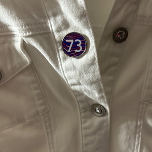 Puede incluir: Un pin redondo con el número 73 en blanco, sobre un patrón ondulado azul y rojo. El pin está sujeto a una chaqueta vaquera blanca, mostrando su diseño y colocación en la ropa.