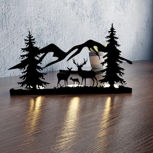 May include: Black metal silhouette of a mountain scene with two pine trees, a deer family, and a mountain range. The silhouette is lit from below with warm white lights.