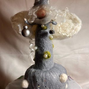 Könnte beinhalten: Ein skurriles Wichtel-Figürchen aus Filz mit grauem Körper, weißem Bart und einer gepunkteten Zipfelmütze. Der Wichtel hält kleine silberne Ornamente und ist mit Perlen und dekorativen Akzenten verziert. Eine einzigartige Weihnachtsdekoration.