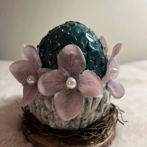 Op de afbeelding: Een decoratief ei met een getextureerd, donkergroen oppervlak, versierd met lavendelkleurige stoffen bloemen en imitatieparels. Het ei rust op een grijze gebreide basis, omgeven door een nest van twijgen en een houten schijf.