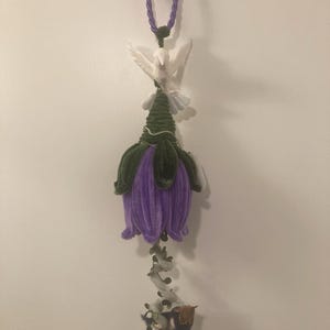 Op de afbeelding: Een hangende bloemvormige ornament in paars en groen met een witte duif en een klein vogeltje dat eronder hangt.