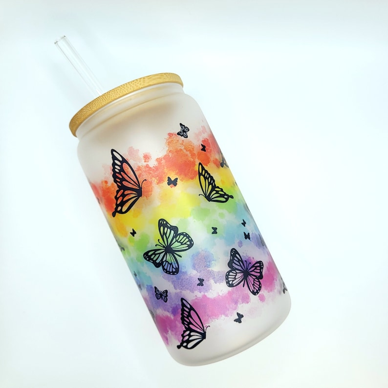 Milchglas mit Bambusdeckel und Glasstrohalm mit Schmetterlingen in Schwarz und im Hintergrund regenbogenfarbende Wasserflecken.