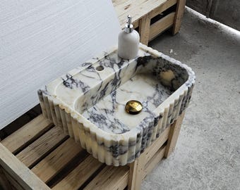 May include: A rectangular marble sink featuring a white base with prominent dark grey and black veining. The exterior of the basin has a vertical fluted texture. A light-colored soap dispenser and a circular gold-toned drain cover are included.