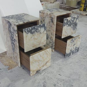 May include: Two marble-patterned nightstands with two drawers each. The drawers are open, revealing a wooden interior. The marble has a mix of white, beige, and gray tones. The nightstands are rectangular and sit on a concrete surface.