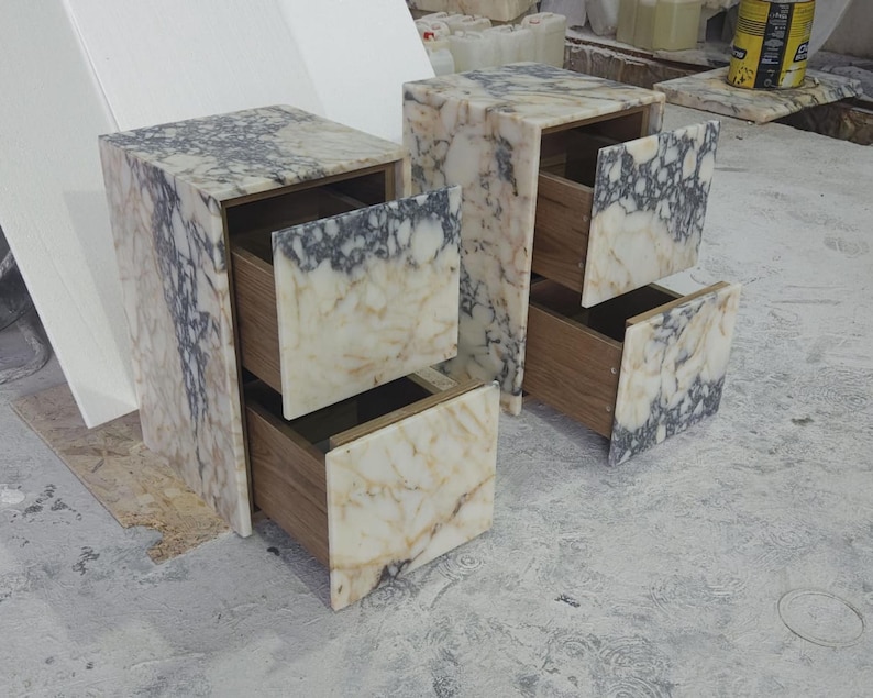 May include: Two marble-patterned nightstands with two drawers each. The drawers are open, revealing a wooden interior. The marble has a mix of white, beige, and gray tones. The nightstands are rectangular and sit on a concrete surface.
