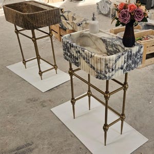 May include: Two stone washbasins with brass-coloured metal stands. One basin is brown, the other is white and grey. The stands have a decorative design. A vase of flowers and a soap dispenser are on the countertop.