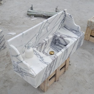 Op de afbeelding: Een wastafel van wit marmer met opvallende donkergrijze aders, een hoge achterwand en gebogen zijpanelen. De waskom is voorzien van een goudkleurige afvoerplug en een witte zeepdispenser staat op het vlakke gedeelte naast de wasbak.