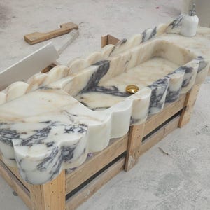 Puede incluir: Un lavabo rectangular de m&aacute;rmol con borde festoneado y vetas en crema, blanco y gris oscuro. La cubeta incluye un desag&uuml;e de tono dorado y un dispensador de jab&oacute;n blanco se encuentra sobre la superficie plana de la encimera de piedra.