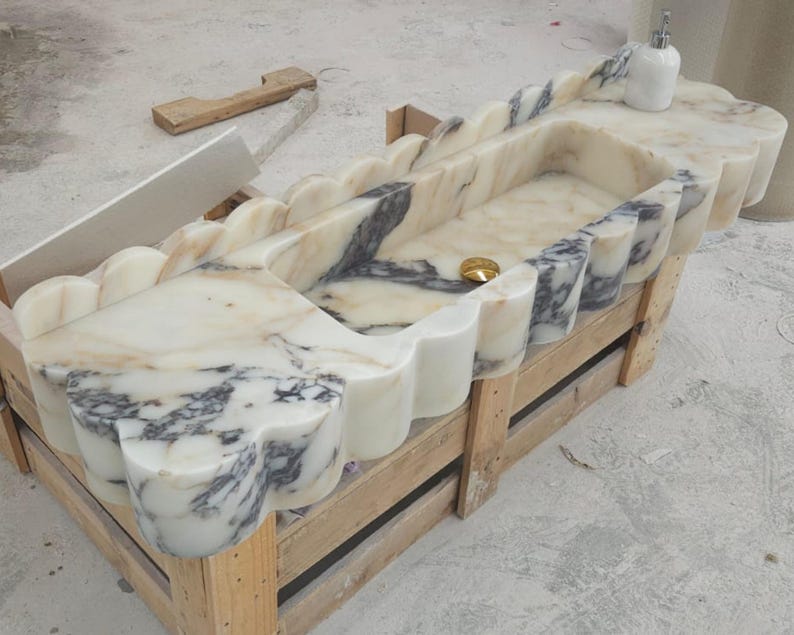 Puede incluir: Un lavabo rectangular de m&aacute;rmol con borde festoneado y vetas en crema, blanco y gris oscuro. La cubeta incluye un desag&uuml;e de tono dorado y un dispensador de jab&oacute;n blanco se encuentra sobre la superficie plana de la encimera de piedra.