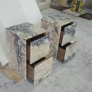 May include: Two marble nightstands with two drawers each. The nightstands have a cream and gray veined pattern. The drawers are open, revealing the wooden interior. The nightstands are rectangular and have a modern design.