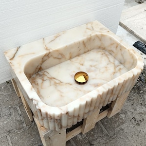 May include: A rectangular marble sink with a scalloped edge design. The sink is white with brown veining and a gold-colored drain cover. It is set on a wooden support structure.