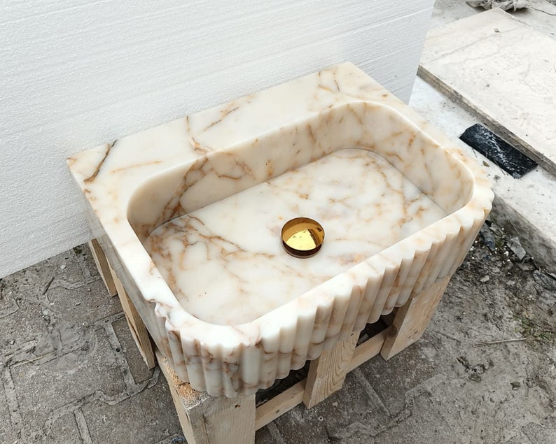 May include: A rectangular marble sink with a scalloped edge design. The sink is white with brown veining and a gold-colored drain cover. It is set on a wooden support structure.